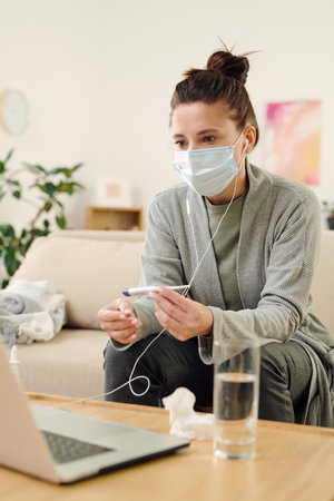 Sick woman in earphones and protective mask looking at doctor on laptop display
