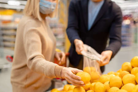 Mature couple in casualwear and protective masks choosing fresh ripe mandarines in supermarket while woman taking one from displayの写真素材