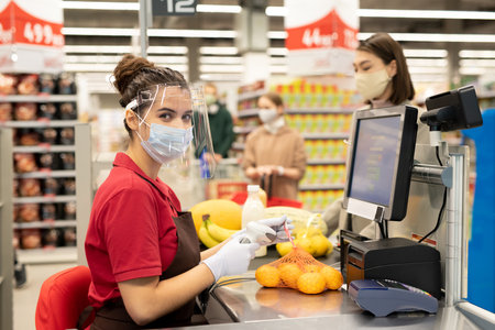 Young cashier in protective gloves, mask and screen scanning pack of orangesの写真素材