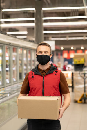 Young courier in uniform and protective mask holding box with fresh productsの写真素材