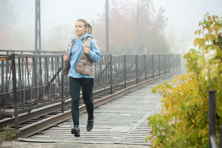 Young pretty sportswoman in activewear jogging along bridge and yellow treesの写真素材