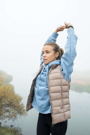 Young sportswoman in black leggins, blue hoodie and grey jacket doing exercise