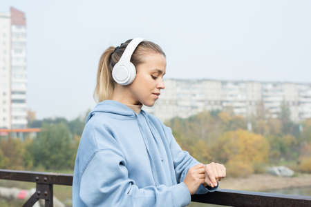 Young serious blond sportswoman in headphones and hoodie looking at wristwatch