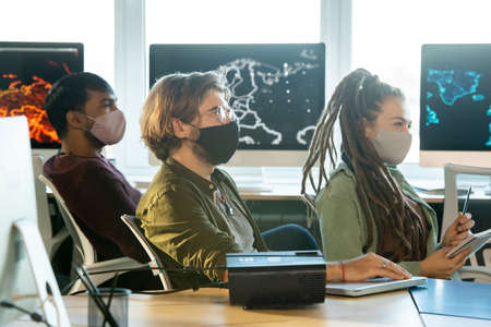 Group of students in protective masks sitting by desks and listening to coachの写真素材