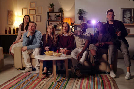Group of young intercultural friends sitting on long couch and watching movie