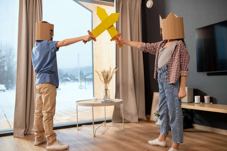 Two little siblings in casualwear and cardboard helmets playing with toy swordsの写真素材