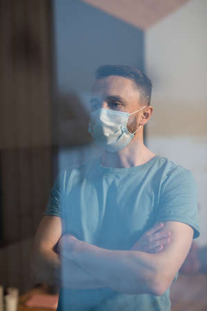 Young man in protective mask looking through window while staying at homeの写真素材