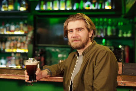Young smiling bearded man in casualwear having glass of beer against bar