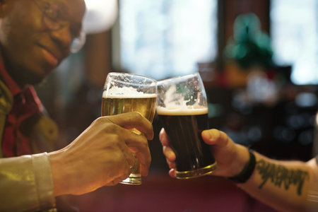 Hands of two young intercultural men clinking with glasses of beer in the pub
