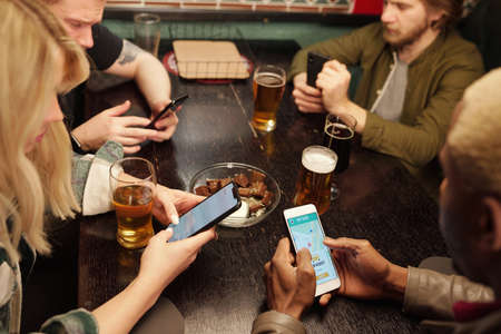 Group of young intercultural friends having beer and scrolling in mobile gadgetsの写真素材