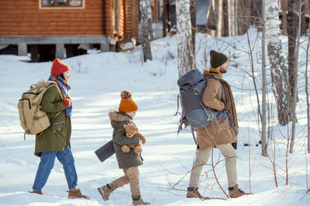 Young man with rucksack, his wife and daughter moving towards country house
