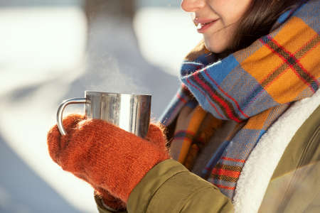 Hands of young female in warm jacket, scarf and knitted mittens holding drink