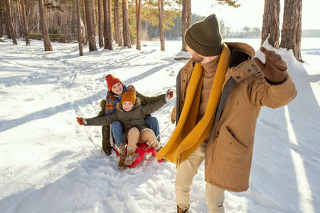 Cheerful young man in warm winterwear pulling sledge with his wife and daughterの写真素材
