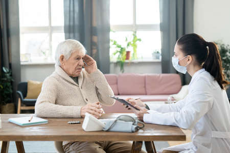 Sick senior man touching head and looking at young female doctor in whitecoatの写真素材