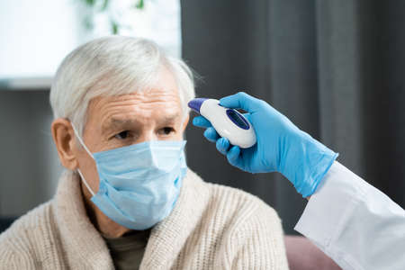 Sick old man in mask sitting in front of doctor using contactless thermometer