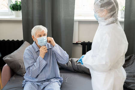 Young female doctor in gloves and coveralls standing in front of sick senior man