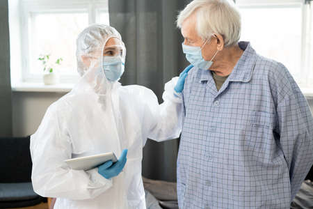 Young female doctor with tablet keeping hand on shoulder of sick senior patientの写真素材