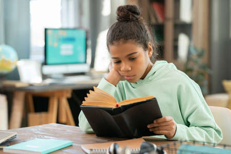 Serious elementary schoolgirl in casualwear reading book by table in living-room