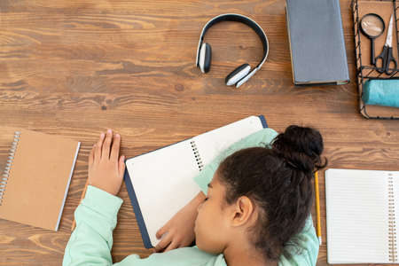 Overview of tired mixed-race schoolgirl keeping her head on open copybook