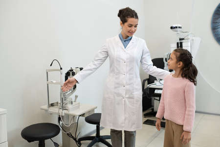 Young brunette ophthalmologist in whitecoat showing little girl new equipment