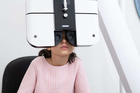 Cute elementary schoolgirl sitting in front of ophthalmological equipment