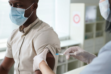Gloved hand of nurse in uniform and protective mask vaccinating African guyの写真素材