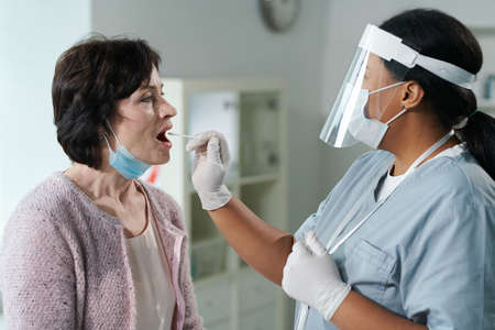 Woman with open mouth sitting in front of gloved nurse testing her for covid