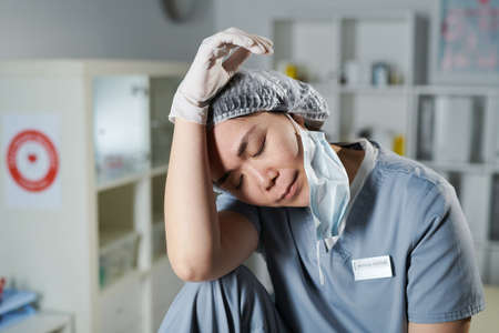 Young tired Asian nurse sitting in front of camera and touching her head while keeping eyes closedの写真素材