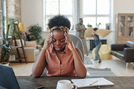Young tired African woman touching head while sitting by table in front of laptop during networkの写真素材