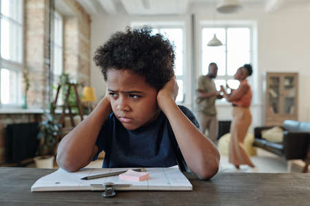 Mixed-race annoyed schoolboy covering ears by hands while sitting by table and trying to do homeworkの写真素材