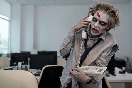 Zombie businessman with phone receiver by ear standing by workplace in front of cameraの写真素材