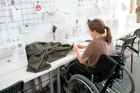 Young disable female in casualwear sitting in wheelchair by workplace inside contemporary workshopの写真素材