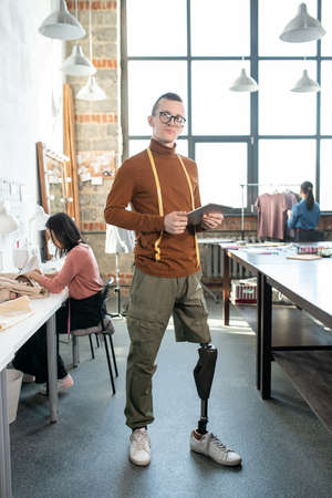 Young fashion designer or tailor with prosthetic leg using tablet by workplace against colleaguesの写真素材