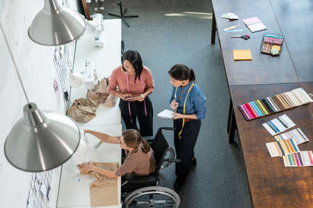 Two young fashion designers standing by seamtsress in wheelchair while looking for creative ideasの写真素材