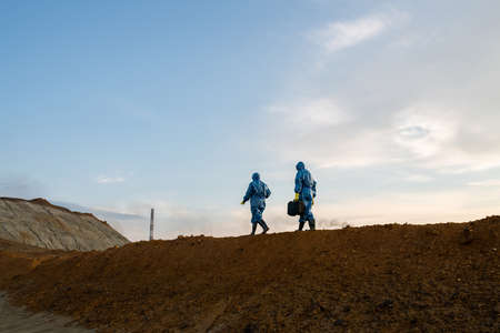 Figures of two contemporary ecologists in protective coveralls moving on top of hill with dirty soil...
