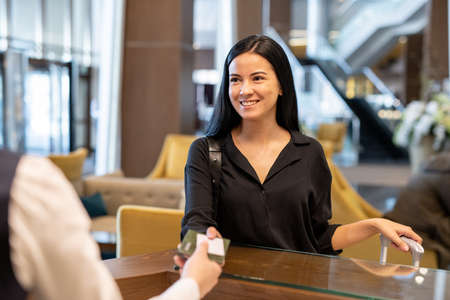 Happy young businesswoman or female traveler with luggage passing her documents to hotel receptionist over counter before registrationの写真素材