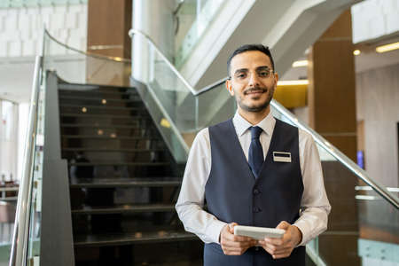 Happy young elegant worker of contemporary hotel using tablet while standing against staircase and looking at you inside loungeの写真素材