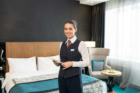Happy young female worker of luxurious hotel in uniform standing in front of camera against bed and using touchpad while looking at youの写真素材