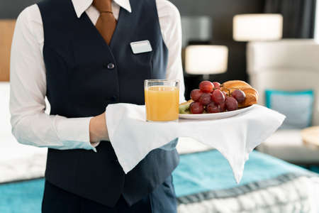 Midsection of young elegant female worker of hotel holding tray with breakfast while standing in front of camera against bedの写真素材