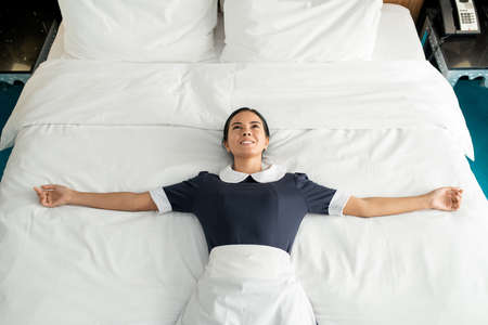 Young cheerful and relaxed chambermaid with toothy smile lying on clean and changed bed with her arms outstretched and enjoying the momentの写真素材