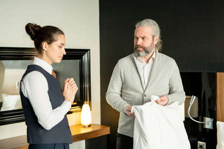 Dissatisfied mature businessman complaining young female manager of hotel about stain on pillow while looking at her with angry expressionの写真素材