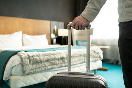 Hand of male traveler or businessman in casualwear holding by handle of his suitcase while standing in front of done bed and going to relaxの写真素材