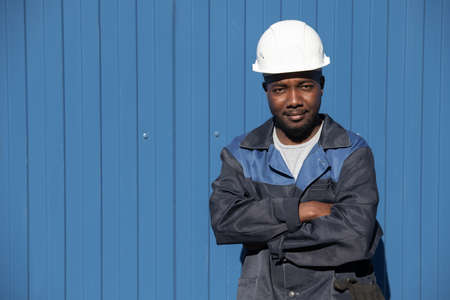 Happy young factory worker of African ethnicity in workwear and hardhat crossing his arms by chest while standing in front of camera outdoorsの写真素材