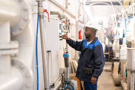 Young African male worker in overalls and hardhat standing in front of huge industrial machine and...