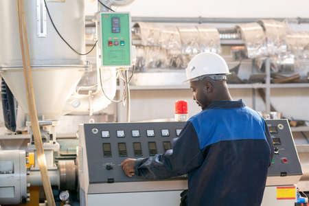 Back view of young African male worker in overalls and hardhat standing in front of control panel of huge industrial machine and turning knobの写真素材