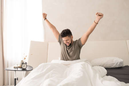Happy young man stretching his arms upwards while sitting on large comfortable double bed under white blanket in the morningの写真素材