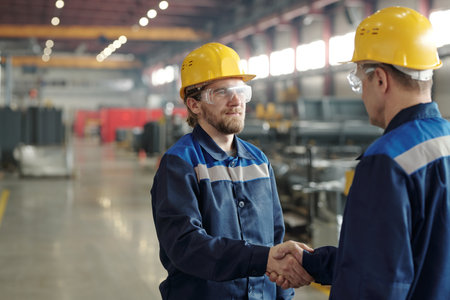 Two male engineers in protective hardhats and workwear shaking hands of one another against large...