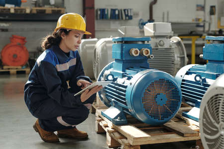 Young serious female engineer in protective helmet and workwear squatting by new industrial...