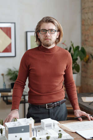 Young elegant male architect in smart casualwear standing by workplace with model of new house and...