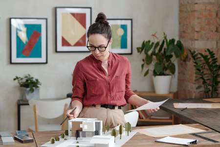 Young elegant female architect with paper and pen standing by table with model of new house and yard...